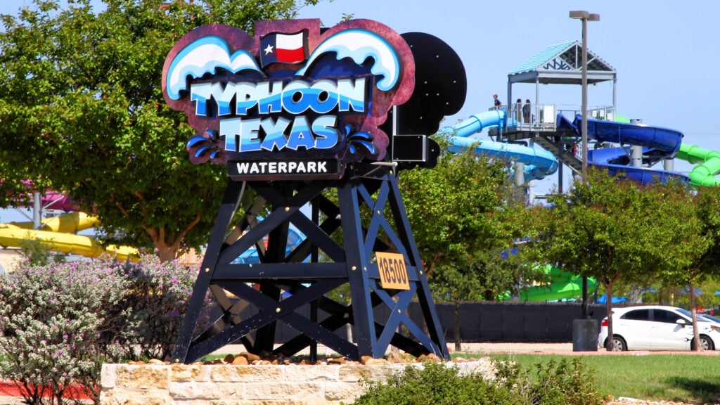 Entrance sign of Typhoon Texas Waterpark with colorful slides and a tower visible in the background, surrounded by trees and a parked car.