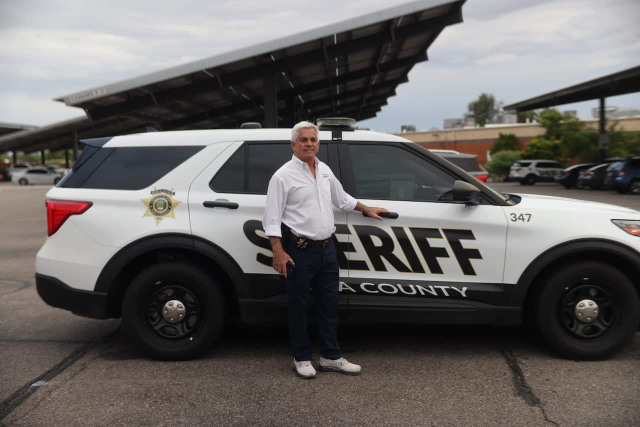 A man in a white shirt and dark pants stands before a white and black four wheel driver with Sheriff written on the door.