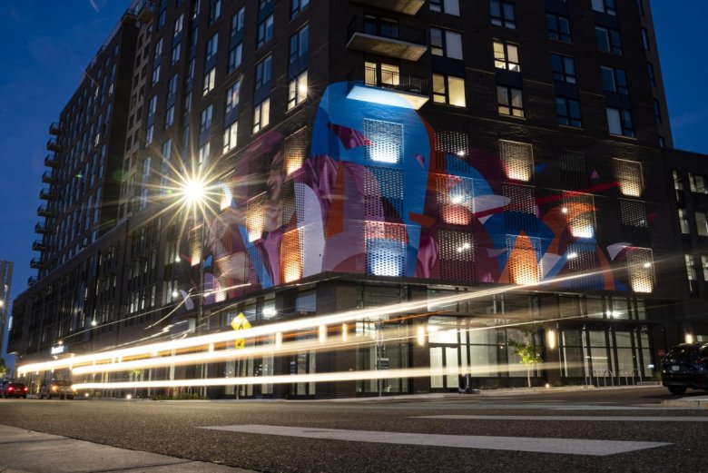 A building with a colorful mural is illuminated at dusk, with light trails from passing vehicles visible on the street.