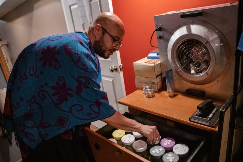 Benji “Dez” Dezaval wearing a colorful shawl is organizing jars in a drawer next to a large machine in a room.