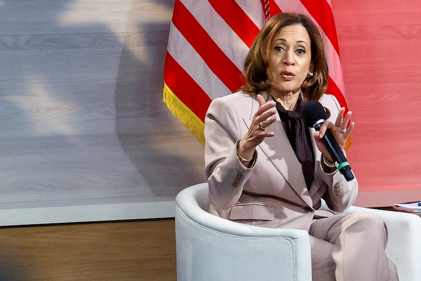 Kamala Harris sits in a chair in front of an American flag and holds a microphone as she speaks.