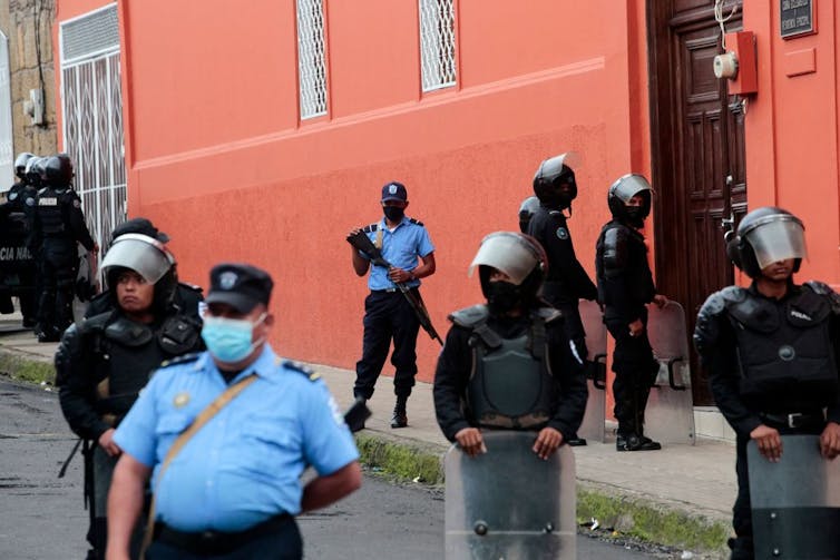 Officers in helmets and shields, one of whom holds a large gun, stand outside a building with orange walls.