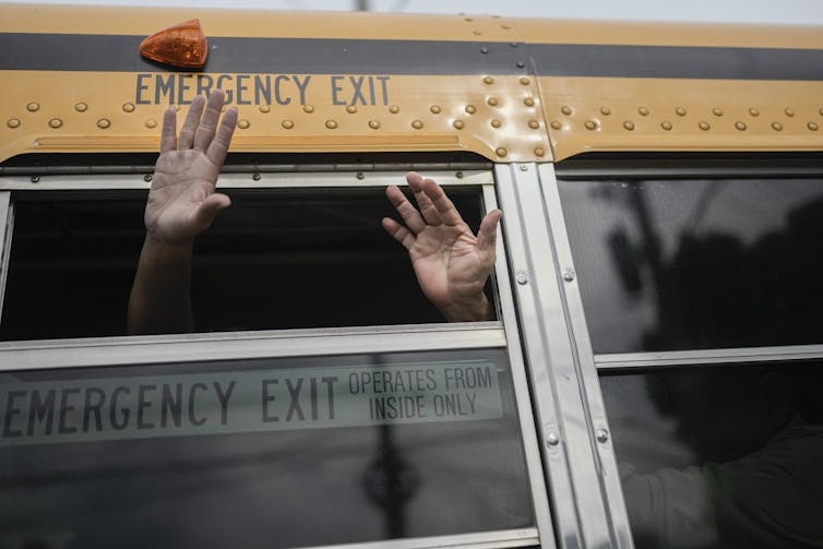 Two hands wave from a half-opened window on a yellow school bus.