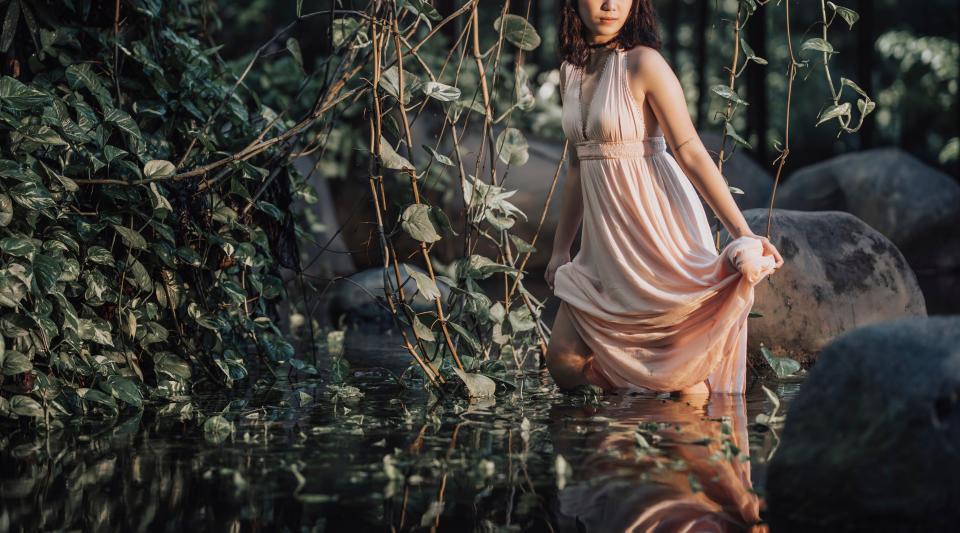 A woman in an elegant, flowing dress kneels in a lush, forest-like setting with water and greenery around her