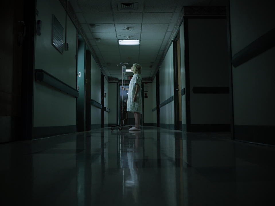 A person in a hospital gown and cap stands alone in a dimly lit hospital hallway, facing to the right while holding an IV pole