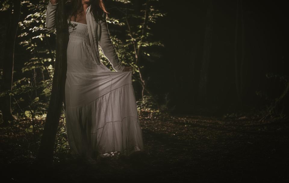 A woman in a flowing dress stands in a dark forest, illuminated by a light source, creating a mysterious and ethereal atmosphere