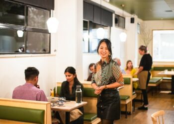 A woman standing in the middle of a restaurant's white dining room with several diners eating at booths