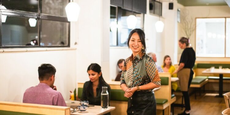 A woman standing in the middle of a restaurant's white dining room with several diners eating at booths