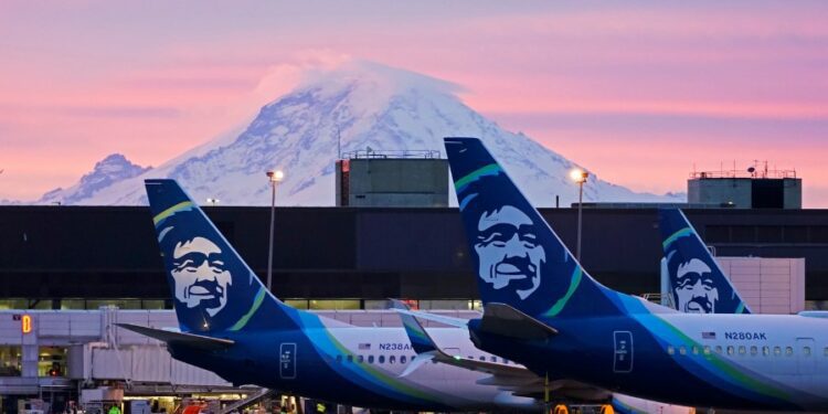 Image: Alaska Airlines planes are shown parked at gates with Mount Rainier in the background at sun...
