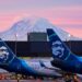 Image: Alaska Airlines planes are shown parked at gates with Mount Rainier in the background at sun...