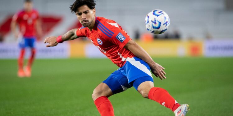 Victor Davila flicks the ball on the pitch, in his red shirt and blue shorts, ahead of the Chile vs Bolivia live stream.