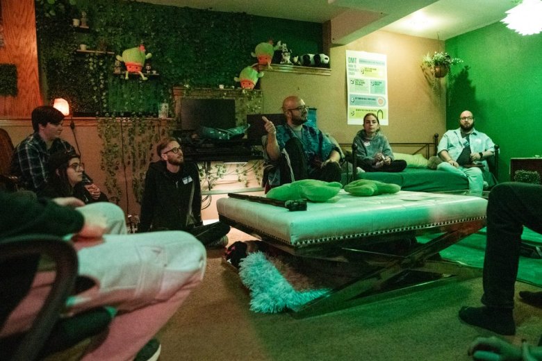 A group of people sitting in a room with green lighting, plants, and posters on the walls. Some are seated on chairs, while others sit on the floor around a central table.