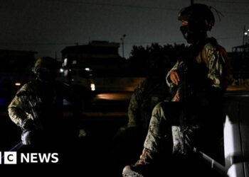 Two people in military fatigues sit in the bed of a truck in a street enveloped in darkness