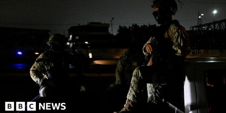 Two people in military fatigues sit in the bed of a truck in a street enveloped in darkness