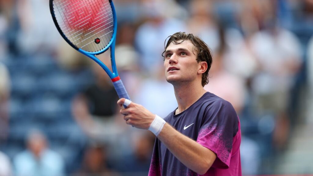 Jack Draper raises his racket at the US Open tennis, wearing a purple Nike t-shirt.