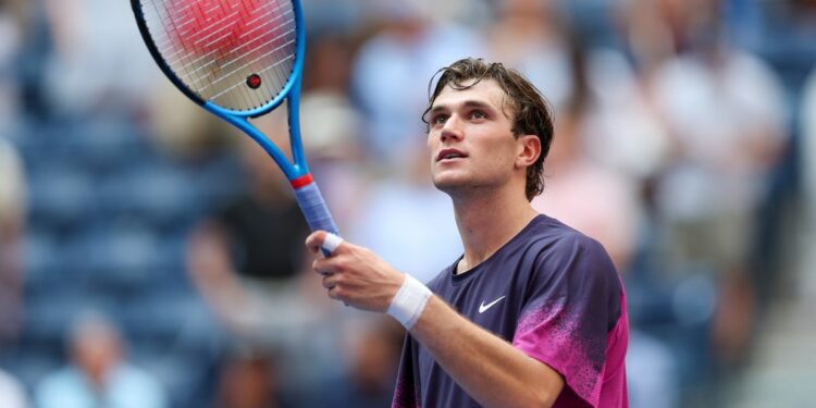 Jack Draper raises his racket at the US Open tennis, wearing a purple Nike t-shirt.