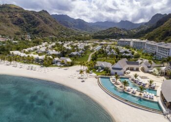 Aerial view of beach and large resort, with curved pool on Buccament Bay and green mountains in background