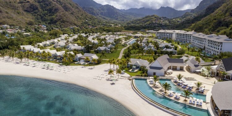 Aerial view of beach and large resort, with curved pool on Buccament Bay and green mountains in background