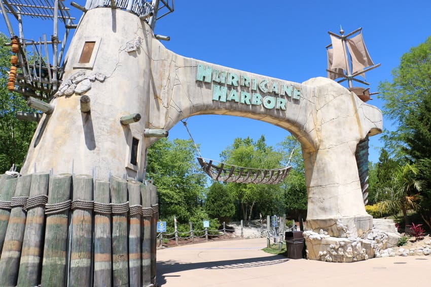 An entrance arch to Hurricane Harbor water park, designed to look like a weathered shipwreck. Surrounded by trees, with clear blue sky in the background.