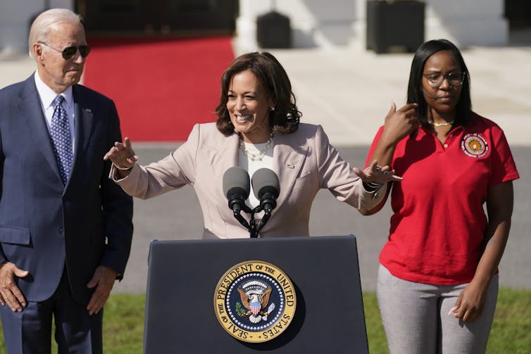 A dark-haired woman stands behind the presidential lectern in front of the White House. An older man in aviator sunglasses stands to her right and a young Black woman stands to her left.