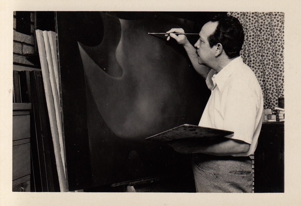 A black and white photo of a middle-aged Rafael Soriano painting a dark abstract canvas in the early 1970s.