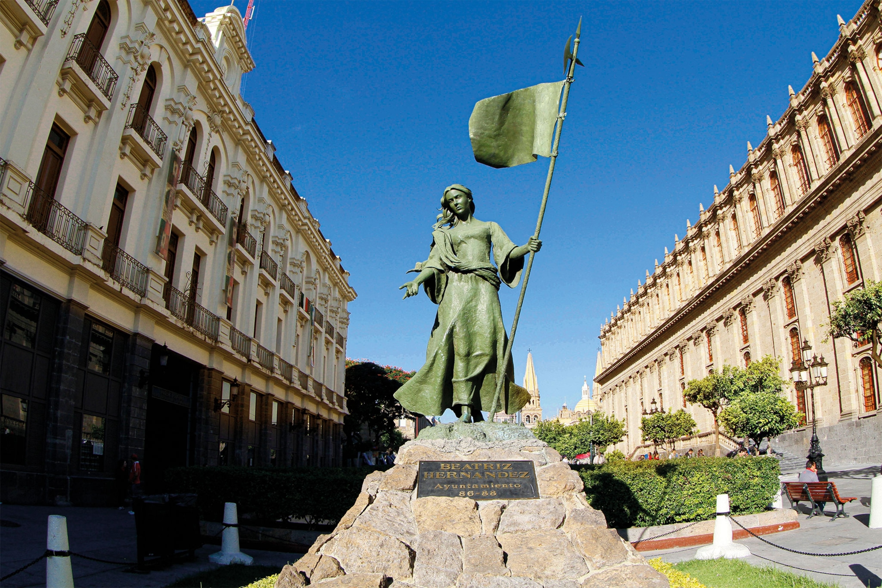 A bronze statue of Beatriz Hernández holding a flag stands near a city plaza