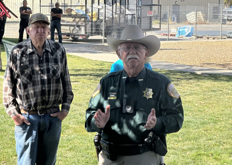 The sheriff, wearing a cowboy hat, speaks outdoors to a crowd of onlookers.