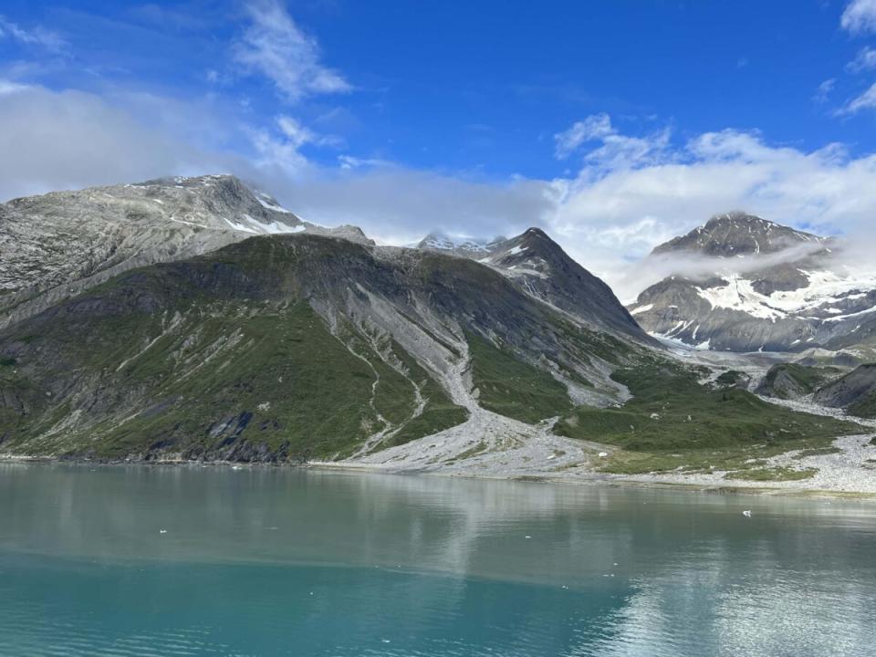 Turquoise waters brush against a green mountain with a snowy mountain in the background in Glacier Bay, AK