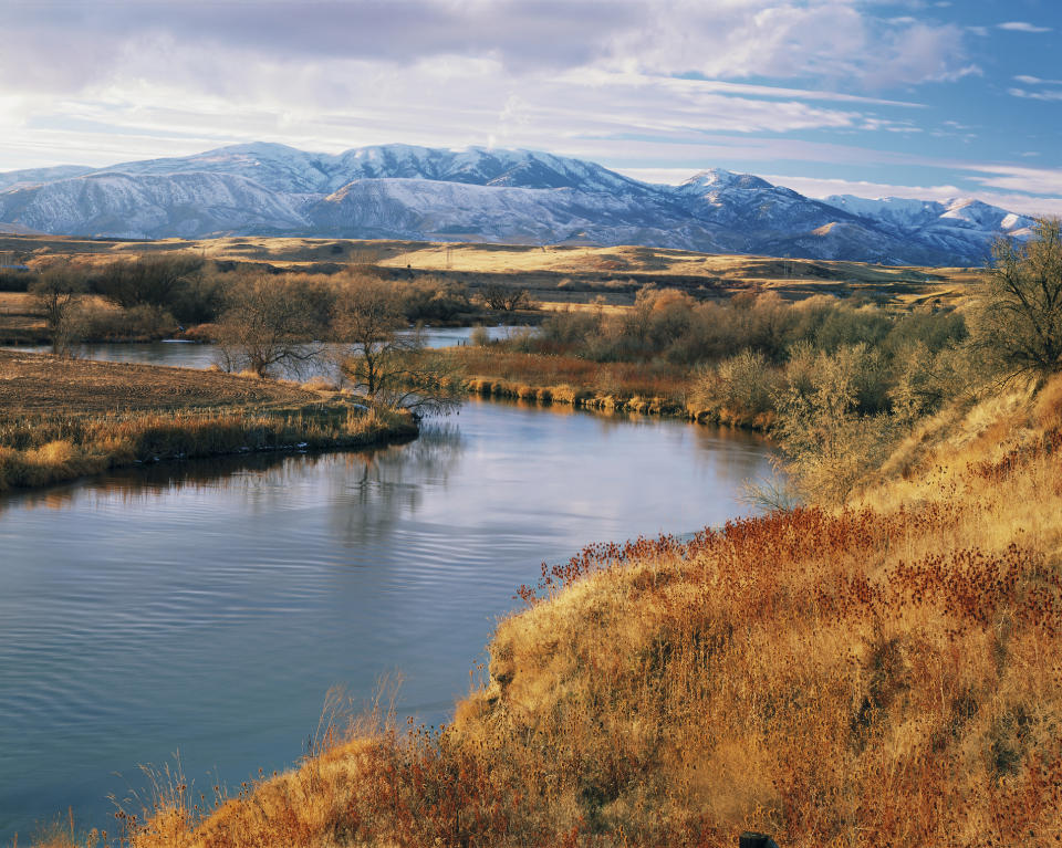 Scenic view of a calm river winding through a landscape of grasslands and leafless trees, with a backdrop of snow-capped mountains