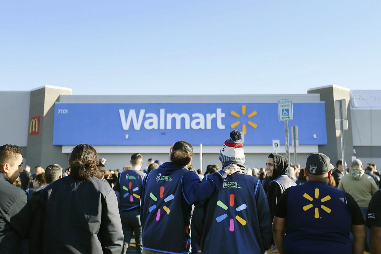 People seen from behind, some wearing blue jackets with a logo, stand facing a building with a blue sign that says Walmart