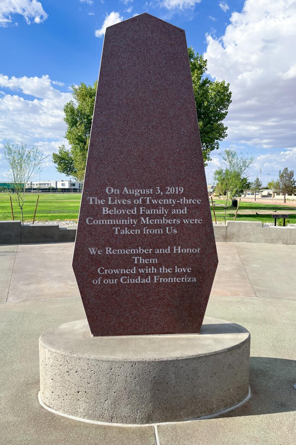 An obelisk-shaped slab sits on a round base on paved ground, with a lawn and trees in the background