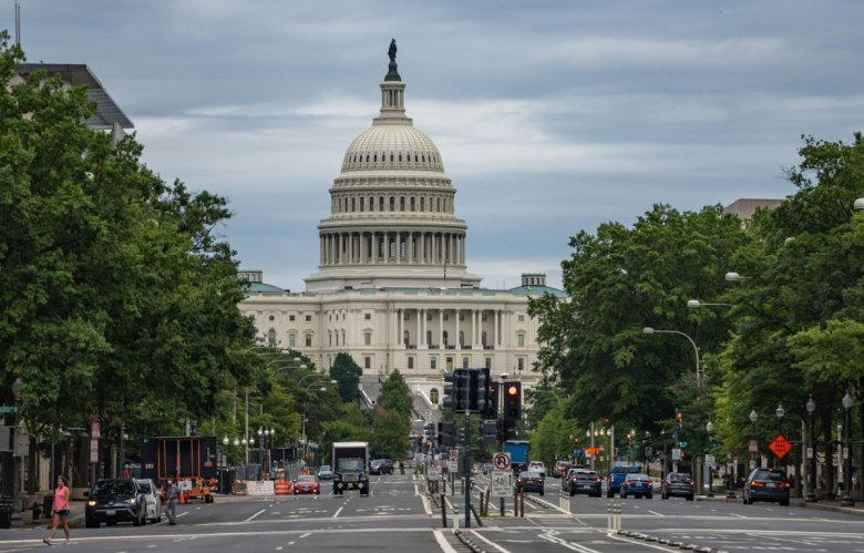 Street view of the U.S. Capitol building with surrounding trees and traffic in Washington, D.C. under a cloudy sky.