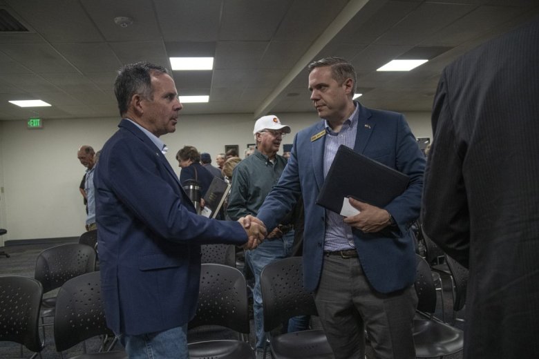 Two men shaking hands in a room with seated and standing people. One holds a folder.