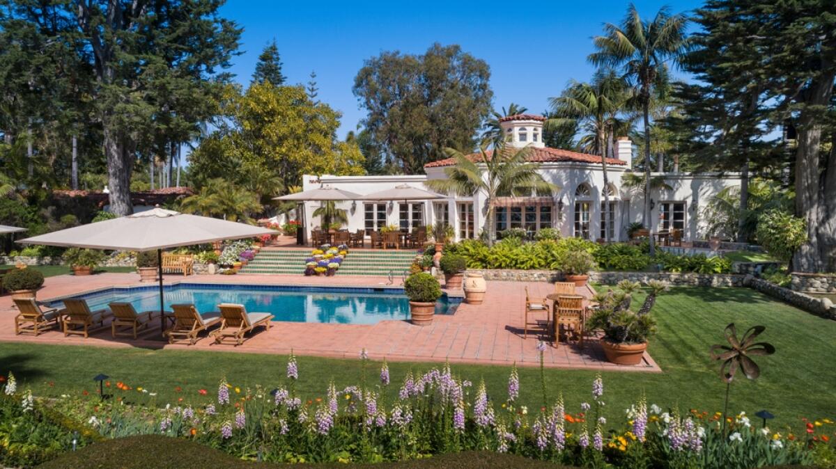 A pool with lounge chairs and an umbrella sits behind a large white home surrounded with mature trees  