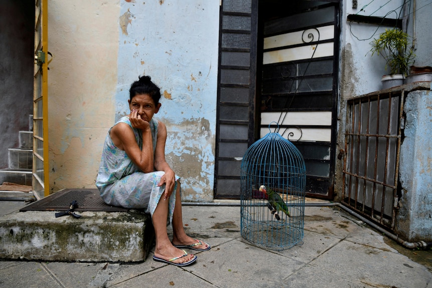 A woman sitting next to a bird in a cage on the street. 