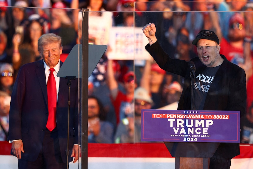 Elon Musk raising his right fist while standing alongside Donald Trump at a rally lectern in front of a crowd of supporters