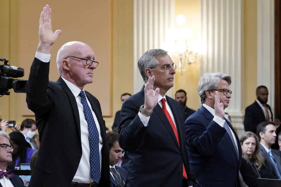 Three men in suits and ties raise their hands in a large room with pale walls.