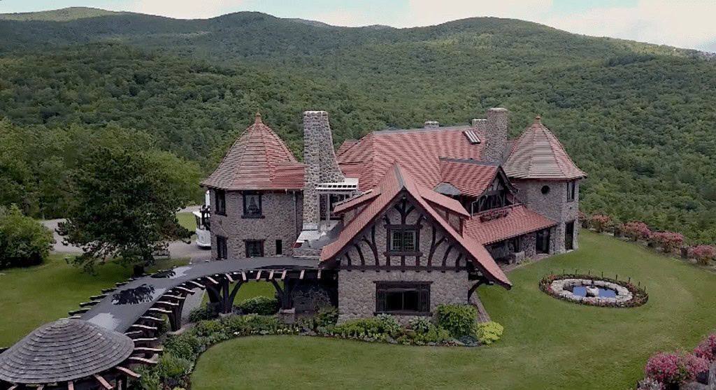 Stone house with red roof and multiple turrets set in a lush, green hillside landscape. Circular driveway and small garden visible.