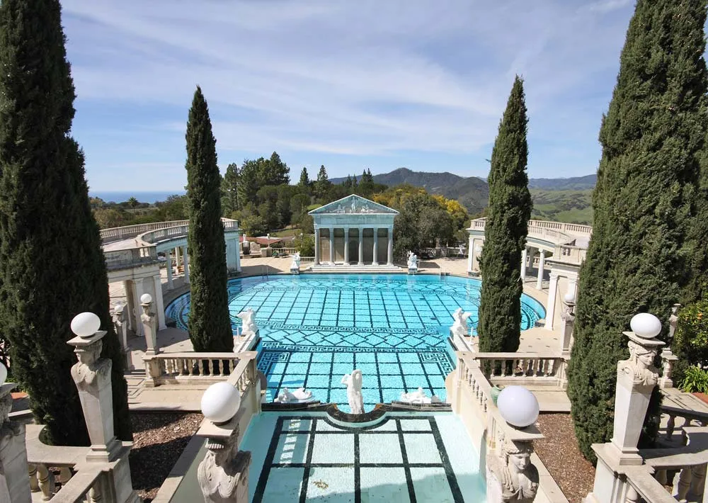 A grand outdoor pool surrounded by tall cypress trees, statues, and classical architecture under a blue sky.