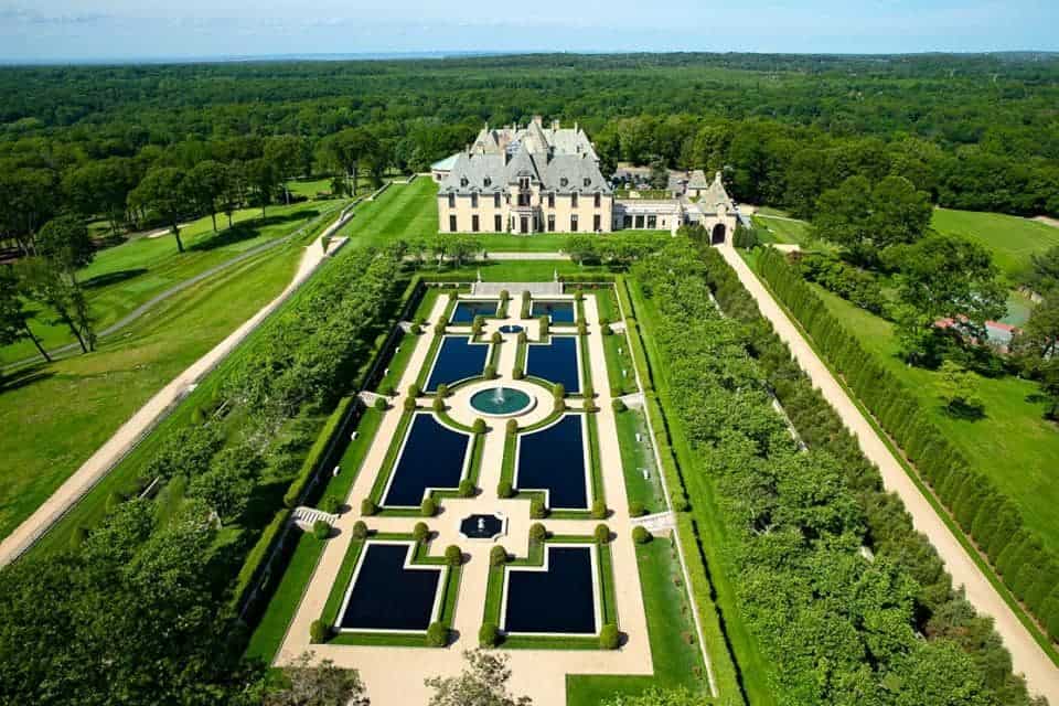 Aerial view of an ornate French-style garden with symmetrical hedges, pathways, ponds, and a central fountain, set in front of a large mansion surrounded by dense greenery.