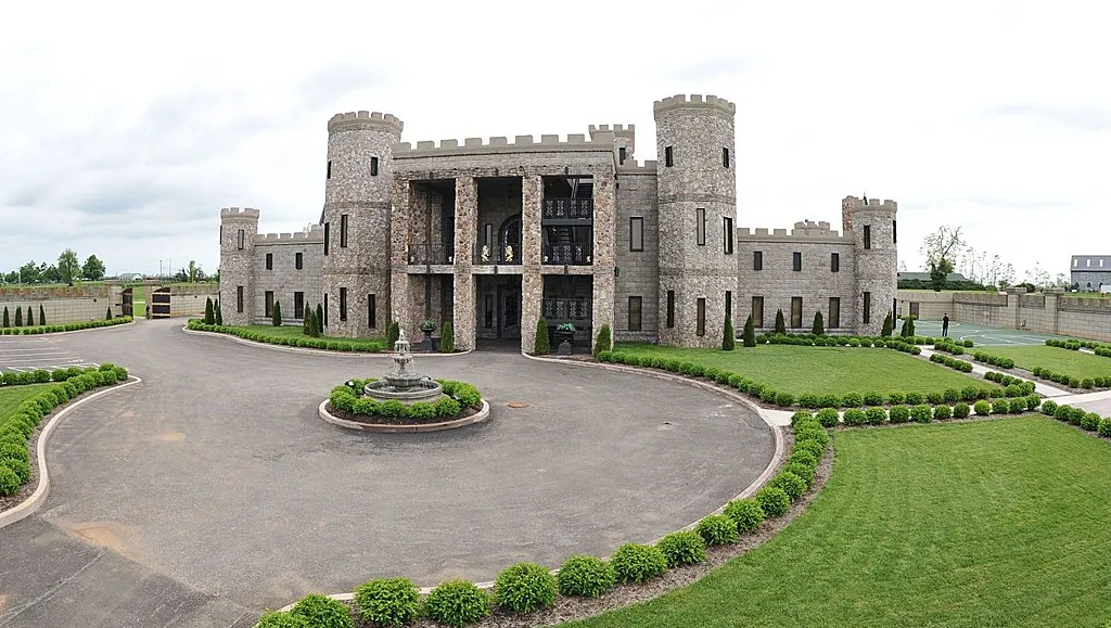 Stone castle with circular driveway, manicured lawns, and a small central fountain under a cloudy sky.