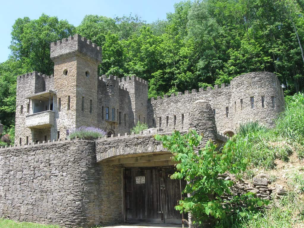 Stone castle with multiple towers, set against a backdrop of lush green trees. Wooden gate at the entrance with a small sign. Bright, clear day.