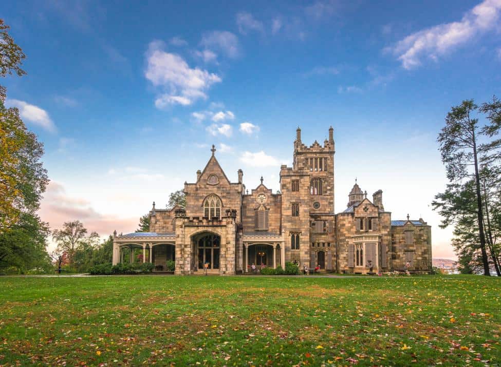 A grand historic stone mansion with multiple towers and arched windows stands surrounded by a green lawn and trees under a partly cloudy sky.