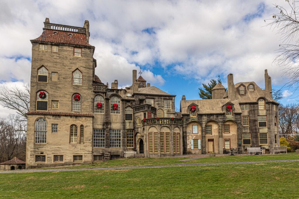 A large, historic stone mansion with multiple towers and red wreaths on windows, set against a partly cloudy sky with a grassy foreground.