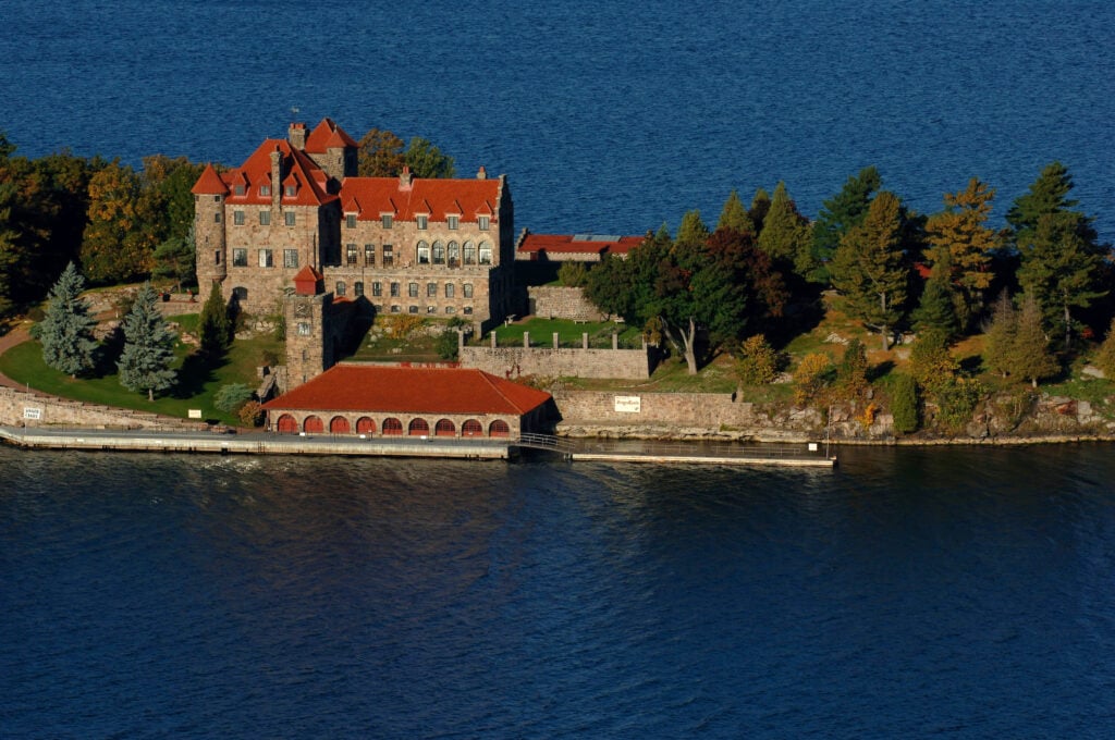 Aerial view of a stone castle with red roofs on an island surrounded by water, bordered by trees and a dock.