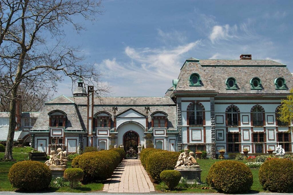 A large, ornate mansion with a slate roof and arched entrance, surrounded by manicured hedges and statues, under a blue sky.