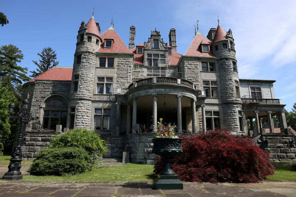 A large, historic stone mansion with multiple turrets and a central portico. The building is surrounded by trees and features a manicured lawn and a decorative urn planter in the foreground.