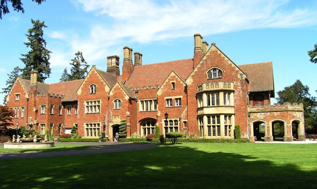 Large, red-brick mansion with multiple chimneys and gabled roofs, surrounded by a manicured lawn and trees under a blue sky.