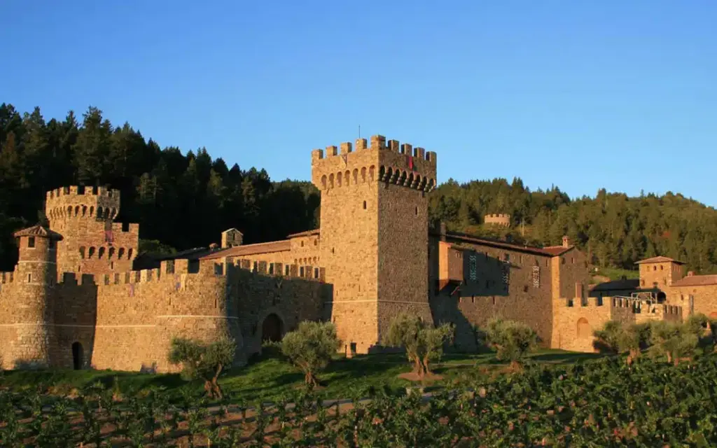 Stone castle with towers, surrounded by vineyards and trees under a clear blue sky.