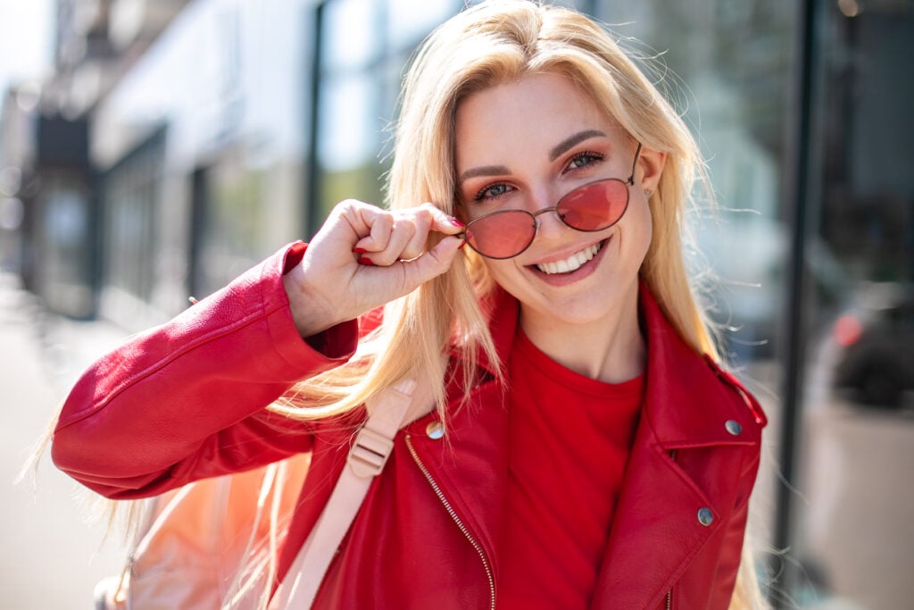 A smiling person with long blonde hair wearing a red jacket and red-tinted sunglasses stands outdoors, holding the sunglasses slightly away from their face.